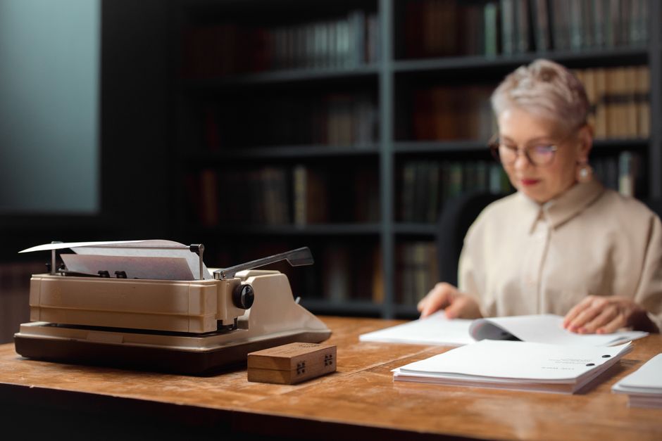 Elderly woman reviewing documents at a desk with a vintage typewriter.